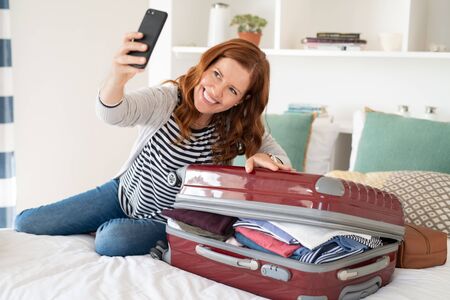 Beautiful mature woman taking selfie with overstuffed suitcase before leaving for a trip. Cheerful middle aged woman sitting on bed using smartphone taking picture with unpacked bag before vacation. Mid woman in video call at home preparing for travel.の写真素材