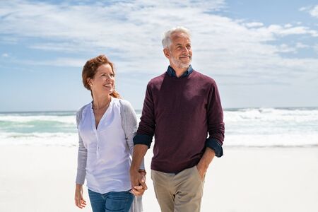 Happy senior couple holding hands at beach in a sunny autumn day. Loving mature couple enjoying their vacation at the sea shore with copy space. Smiling wife and happy old husband walking barefoot on the white sand.の写真素材