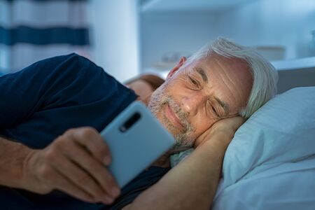 Senior man watching video on smart phone while in bed. Smiling mature man lying on bed using cellphone in the dark at home. Addict man awake using phone for chatting late in night.の写真素材