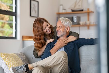 Smiling woman hugging her husband from behind in the living room. Loving mature couple sitting on couch and looking at each other. Mid adult beautiful woman embracing smiling senior at home.の写真素材