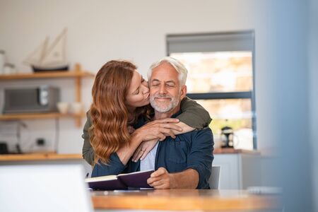 Happy loving woman kissing old man on cheek at home. Beautiful mature wife kissing her husband with beard and closed eyes. Senior couple in love.の写真素材