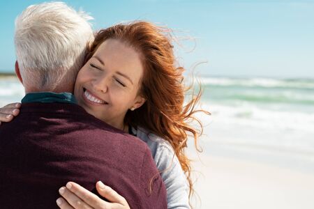 Loving couple embracing at beach with copy space. Portrait of beautiful loving woman hugging senior man at sea with closed eyes. Cheerful couple in love hugging and smiling while relaxing at vacation.の写真素材