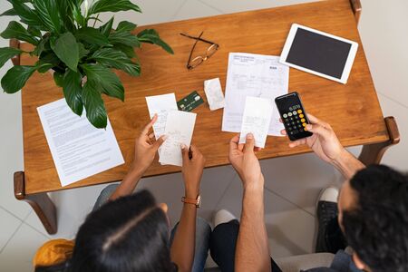 Top view of indian couple checking bills while managing account at home. High angle view of casual man and middle eastern girl using smartphone while looking at invoice and plan the budget to save. Young couple managing home finance and calculating monthly expense.の写真素材