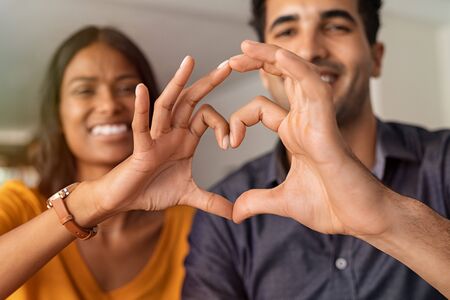Close up of middle eastern couple making heart shape with hands. Beautiful young indian couple in love making heart sign with hands. Portrait of two happy smiling lovers at home.の写真素材