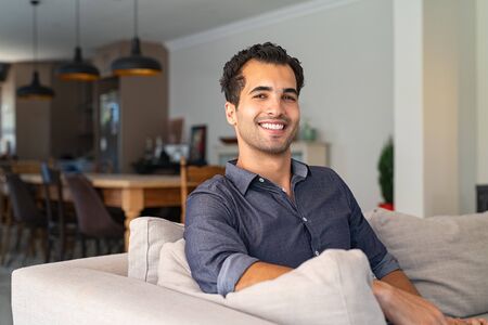 Portrait of happy young middle eastern man sitting on sofa at home. Handsome latin man in casual relaxing on couch in bright living room. Cheerful smiling mixed race guy looking at camera.の写真素材