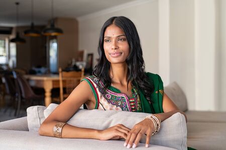 Young pensive indian woman in traditional clothing looking away at home. Beautiful girl wearing sari with a bindi on forehead. Smiling contemplative woman in saree relaxing at home.の写真素材
