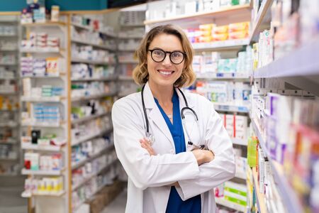 Portrait of mature woman pharmacist at pharmacy wearing labcoat with stethoscope. Happy smiling doctor standing in modern pharmacy drugstore. Friendly young pharmacist owner standing in shop and looking at camera.の写真素材