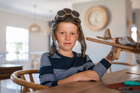 Portrait of little boy wearing pilot helmet playing with wooden airplane at home. School kid playing...