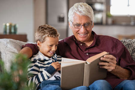 Grandfather and grandson reading a book and smiling. Happy little boy with old grandpa reading story book at home. Senior man tells a fairy tale story to his grandchild while sitting on couch.の写真素材