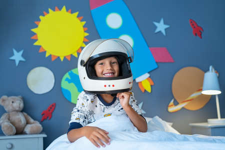 Portrait of happy child wearing astronaut helmet in outer space decorated bedroom. Smiling kid ready to sleep with wall decorated with cardboard planets and rocket. Excited cute boy pretending to be an astronaut looking at camera while sitting on bed before sleep.の写真素材