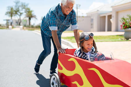 Senior grandfather helping grandson ride on kids toy car. Happy old man pushing car while boy driving on street. Grandparent playing with little boy on gokart.の写真素材