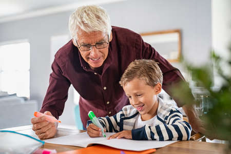 Senior man wearing spectacles helping smart boy complete his homework using highlighter pen. Cheerful school boy preparing project in book with grandfather. Grandson and grandpa drawing together on notebook with sketchpen and smiling.の写真素材