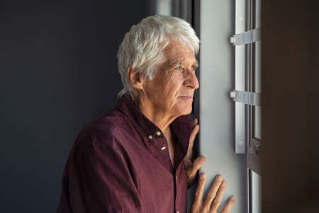 Lonely old man in contemplation near window looking away and thinking. Thoughtful retired man at home looking at view outside window and contemplating. Pensive senior with white hair standing and relaxing copy space.の写真素材