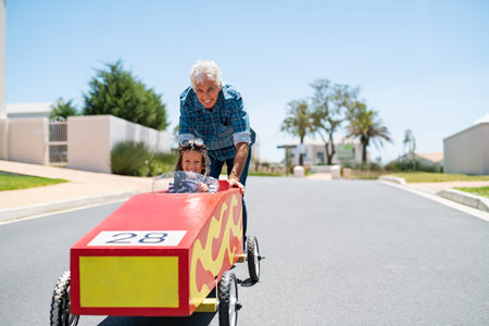 Grandfather helping grandson ride on kids toy car with copy space. Senior man pushing go-kart with boy riding it with pilot helmet. Old grandpa playing with grandchild ride toy car.の写真素材