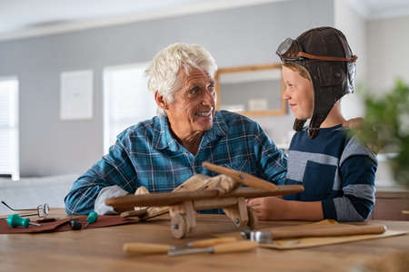 Smiling boy wearing pilot hat and helping making wooden airplane with his grandpa. Happy grandson helping old grandfather making wooden airplane for fun. Playful child wearing aviator helmet enjoying making a plane with senior man.の写真素材