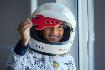 Happy smiling boy wearing an astronaut helmet costume and playing with a spaceship. Child wearing space pajamas and playing with toy rocket near the window. Kid in space dress wearing helmet playing with red toy spaceship at home and dreaming outer space.の写真素材