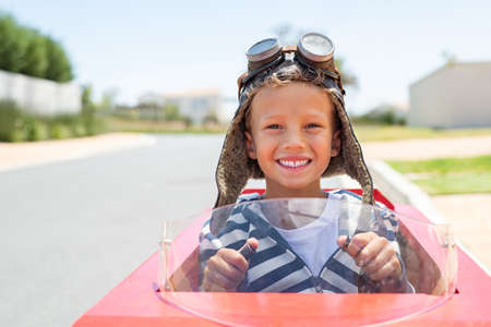 Portrait of little boy riding an handmade toy car on street wearing helmet and pilot goggles. Smiling funny kid rides on a go-kart looking at camera with copy space outdoor. Happy kid sitting in toy car with hands on steering wheel ready for the race.の写真素材