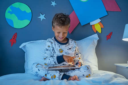 Cheerful boy using digital tablet to search images about solar system and outer space while sitting on bed.の写真素材