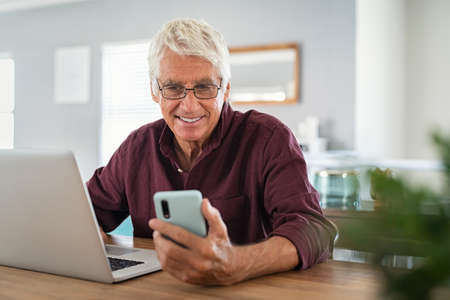 Mature man using smartphone in front of laptop computer. Happy senior businessman working at home while messaging on mobile phone. Happy smiling old man using social media network technology and feeling excited.の写真素材