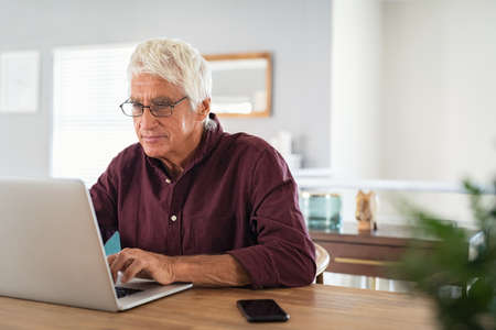 Old man working on laptop at home. Senior man using computer in living room, sitting on chair and looking at screen. Elderly grandfather wearing eyeglasses and working on laptop remotely.の写真素材