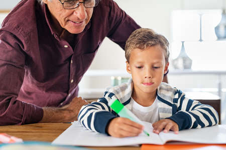 Senior man helping child to complete his homework at home. Focused school boy preparing project on notebook with grandparent. Grandson and grandfather writing exercise on paper.の写真素材