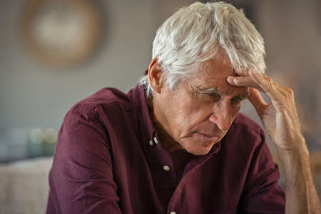 Tired and stressed old man with hand on head sitting at home with headache. Senior retired man at home feeling worried about health problems. Depressed elderly man sitting at home wit head pain suffers from loneliness.の写真素材