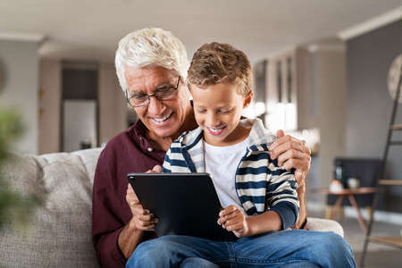 Happy grandfather with grandson relaxing at home while using digital tablet. Playful senior man with child at home playing on digital tablet on sofa. Old grandparent wearing spectacles watching videos with boy sitting on his lap.の写真素材