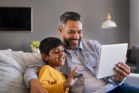 Indian father and smiling son sitting on couch using digital tablet at home. Man and boy using...