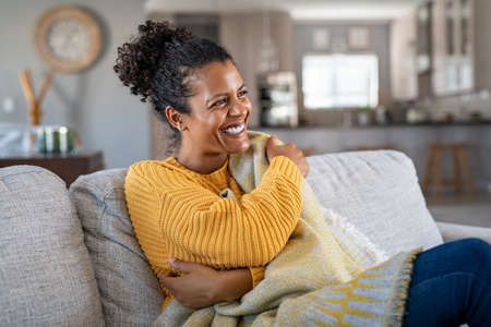 Beautiful black girl sitting on couch wrapped under blanket and laughing. Cheerful african american woman sensitive to the cold relaxing at home on sofa. Carefree and happy mid woman hugging self with warm blanket in winter.の写真素材