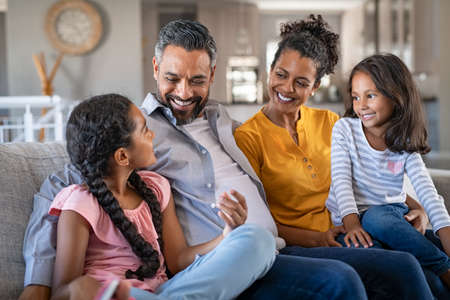 Cheerful multiethnic family at home sitting on sofa listening to daughter. Happy couple with two girls relaxing at home together. Smiling indian parents talking to their daughter at home in the living room.の写真素材