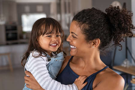 Happy african mother carrying smiling daughter in her arm. Young beautiful woman holding cute little girl in hands. Lovely black mom embracing her child at home.の写真素材