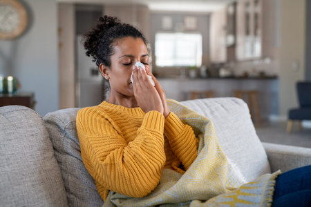 Portrait of young black woman sneezing in to tissue at home. Sick african woman wrapped in blanket...