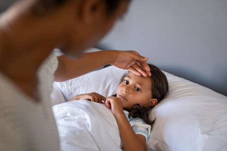 African woman checking temperature with hand of little ill daughter. Mother checking temperature of her sick indian girl. Sick child lying on bed under blanket with woman checking fever on forehead by hand.の写真素材