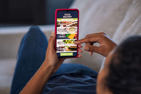 Woman lying on couch using food delivery app on smartphone. Close up of african woman hands holding cellphone and ordering food online. Girl using smart phone to get home delivered take away lunch.の写真素材