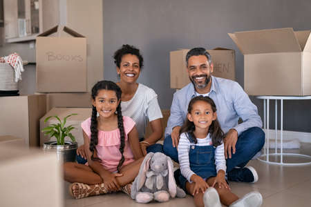 Portrait of smiling ethnic family with two children having fun at new home. Young parents with two daughters in their new house with cardboard boxes looking at camera. Beautiful little girls sitting on floor with african mother and middle eastern dad after relocation.の写真素材