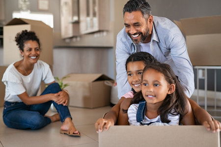 Happy children enjoying cardboard ride with indian father in new home. Cheerful daughters sitting in...