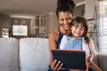 Black young mother and little daughter using digital tablet on couch. Portrait of happy african woman and her cute little girl sitting on sofa together and doing video call on digital tablet. Smiling mom with her child sitting on her laps watching cartoon, copy space.の写真素材
