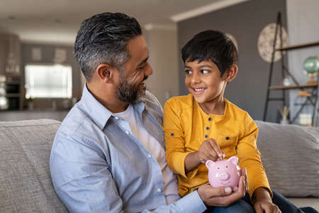 Indian father and smiling son putting coin into piggy bank. Smiling boy sitting on father lap saving money in piggybank. Middle eastern dad teaching son to save money while putting coin in piggybank at home.の写真素材