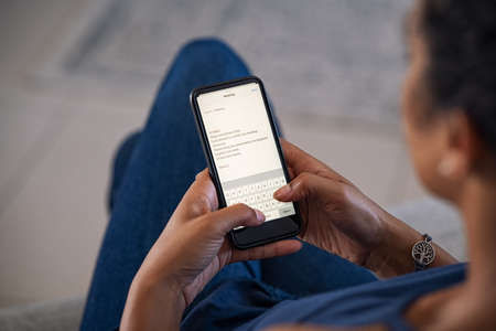 Close up of african woman hands using mobile phone typing an email. Back view of girl writing email on smart phone and replying while working from home. Top view of businesswoman using smartphone while working from home.の写真素材