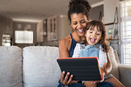 Young black mother and smiling daughter playing on digital tablet at home with copy space. Young african american woman with cheerful and excited little girl using digital tablet while relaxing on couch at home.の写真素材