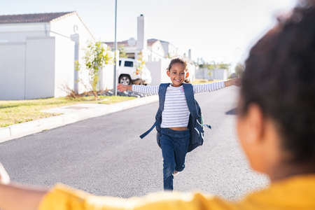 Happy cute little girl running with outstretched arms towards mother outside the house. Young indian schoolgirl feeling happy after first lesson at primary school. Elementary student kid running in the driveway into hands of african mother to hug her after school.の写真素材