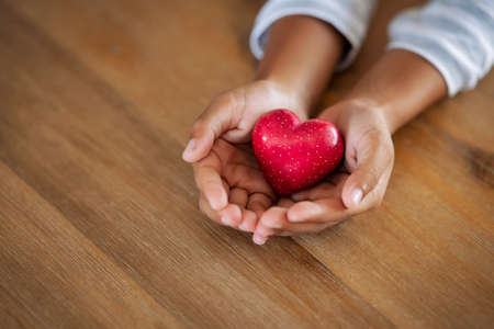 Close up of girl hands holding a red heart shape on wooden table with copy space. Top view of little cute kid hands showing small red heart. High angle view of child holding a heart-shaped stone at home: love, insurance and organ donor concept.の写真素材