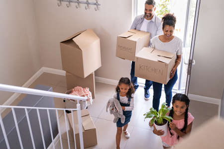 Ethnic family with two children carrying boxes and plant in new home on moving day. High angle view of happy smiling daughters helping mother and father with cardboard boxes in new house. Top view of excited kids having fun walking up stairs running to their rooms while parents holding boxes.の写真素材