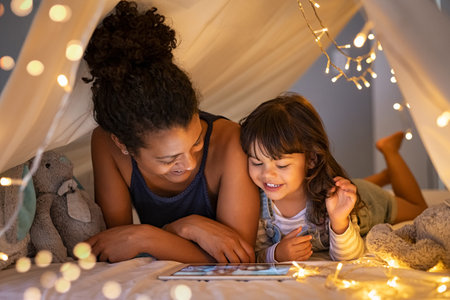 African mother and cute smiling girl using digital tablet while lying in illuminted tent in kid...