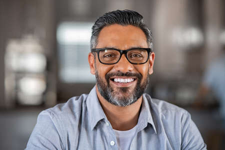 Smiling mature indian man wearing spectacles and looking at camera. Portrait of middle eastern confident businessman at office. Portrait of successful mid entrepreneur feeling satisfied and working from home.の写真素材