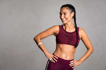 Happy beautiful fit woman laughing and looking away after gym exercise isolated on grey background. Middle aged woman with braides wearing bordeaux sports bra and relaxing on gray wall with copy space. Carefree fitness girl  with big grin resting after workout session: wellness and wellbeing concept.の写真素材
