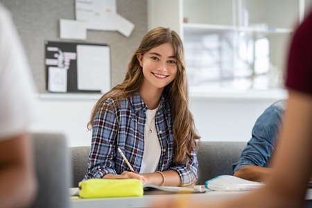 Beautiful university student smiling while studying for exams in classroom. Pretty woman sitting in classroom full of students during class. Portrait of happy young woman writing notes and looking at camera.の写真素材