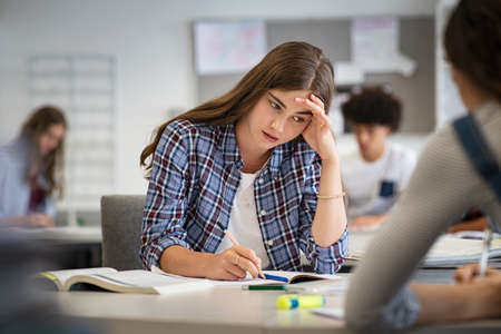 Anxious young woman with hand on head feeling tired while studying at school in classroom. College student suffering from headache in library preparing for test. Portrait of troubled and stressed girl doing exam that doesnât know the answers.の写真素材