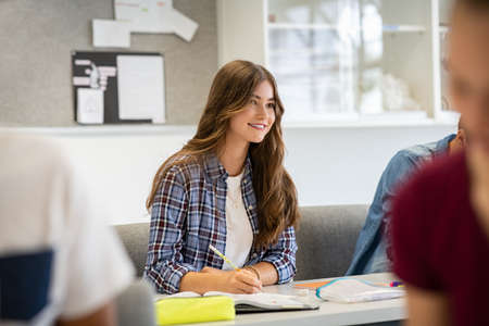 Happy girl in college listening carefully while in lecture. Concentrated student studying and taking notes in a notebook sitting in lecture during lesson. Smiling high school teen sitting in classroom with other students writing notes and listening to professor.の写真素材