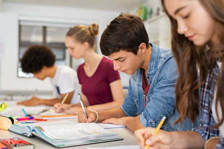 Group of college students studying in classroom writing notes during lesson. Focused guy and girls studying in college library sitting at desk. Group of multiethnic university students doing research sitting in a row.